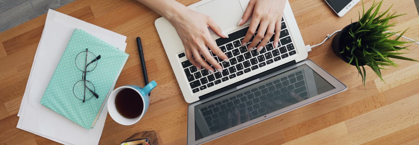 Vogelperspektive auf einen Schreibtisch, auf dem ein Laptop, ein Notizbuch, eine Brille und ein Becher Kaffee stehen. Vor dem Laptop sitzt eine Person, von der man die Hände sieht.